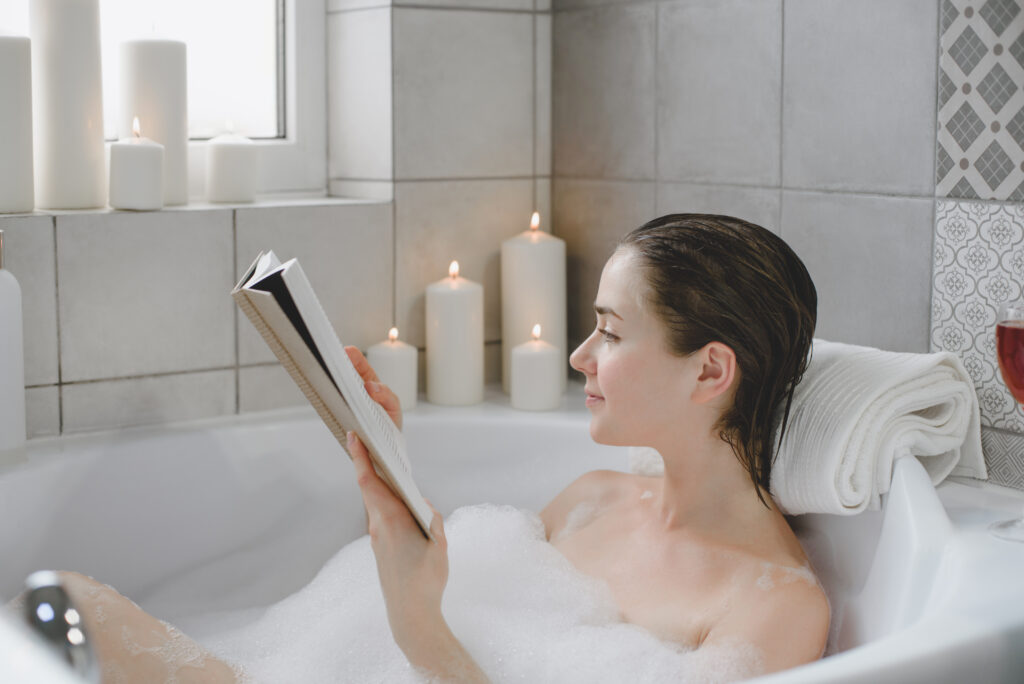 Woman making the most out of her warm bath with a book, bubbles, candles, and wine.